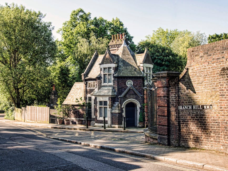 A Victorian-style brick house with ornate architectural details, including a steeply pitched, tiled roof, decorative gables, and tall chimneys, situated behind a brick boundary wall and black wrought-iron gate. The house features white window frames and stone accents contrasting with the red brick facade, and is set against a backdrop of lush green trees and foliage. The street scene includes a paved sidewalk and a quiet road with a double yellow line, indicative of residential UK surroundings. The natural daylight casts soft shadows, highlighting the textures of the brickwork and roof tiles. The location sign reading 'Branch Hill N.W.3' is attached to the brick wall on the right, suggesting an area with private properties that may be serviced for independent waste collection or rubbish removal. Waste Clearance Hampstead offers rubbish disposal services, possibly including on-site clearance of such residential properties, aligning with non-local-authority waste handling options in the area.