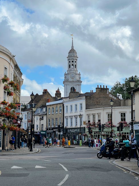 A busy street scene in Hampstead featuring a variety of multi-storey buildings with diverse architectural styles, including brick facades and white-painted exteriors, with shopfronts at ground level and residential or office spaces above. In the foreground, pedestrians are walking along the pavement, some entering or exiting shops, while others are standing or sitting on benches under hanging flower baskets and streetlights decorated with pink and red flowers. To the right, there are a parked motorcycle and a police officer on a scooter. The street appears to be a central part of the village, with a crosswalk and vehicle markings visible on the road. In the background, a prominent historic clock tower with a slender spire and intricate detailing rises above the rooftops, standing against a partly cloudy sky with patches of blue. The scene illustrates a typical high street in Hampstead, with elements that could be associated with independent and private waste collection services like those offered by Waste Clearance Hampstead, in the context of maintaining clutter-free, well-managed town centres.