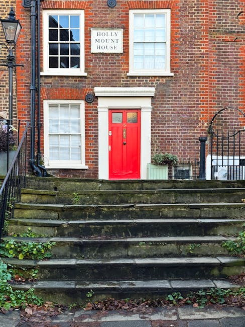 A front view of a brick residential building with a red wooden door, situated above a set of stone steps that are weathered and covered with moss and small green plants. The building has two white-framed sash windows with multiple panes, one located directly above the door and another on the ground floor to the left. A small potted shrub and a green planter are positioned on the right side of the entrance, against the brick wall. A white sign reading 'HOLLY MOUNT HOUSE' is mounted to the upper left of the door. To the right, part of a black wrought iron fence and gate is visible, leading up the steps, partially obscuring an outdoor seating area with a metal table and chair. The scene is outdoors in natural daylight, with an older, slightly weathered appearance of the steps and surrounding vegetation. The context of rubbish removal is implied by the setting, representing a typical residential property that might require on-site clearance or private waste handling services by Waste Clearance Hampstead.