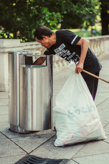 A middle-aged man with dark hair, dressed in a black T-shirt with graphic white text on the front, is seen bending over and using a metal grabber tool to pick up rubbish from a stainless steel litter bin with three compartments on a paved sidewalk. He is holding a large, partially filled white plastic bag in his left hand, which is resting against his thigh. The scene takes place outdoors in a park or street setting, with lush green trees and grass visible in the background, and a stone or concrete balustrade along the side of the pavement. The man appears to be engaged in waste collection, possibly as part of a community cleanup or private rubbish removal service. The lighting indicates it is daytime, with natural sunlight illuminating the scene, emphasizing the clean, reflective surface of the metal bins and the vibrant greenery surrounding the area. This image exemplifies the kind of assistance provided by Waste Clearance Hampstead for alternative waste handling beyond municipal rubbish collection services.