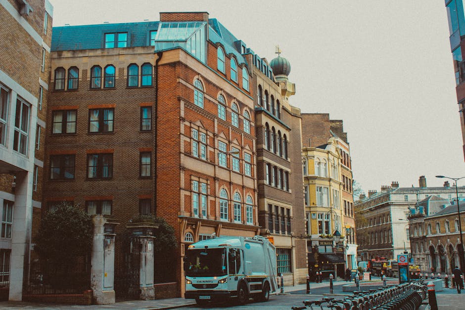 A street scene featuring a modern white waste collection truck parked in front of a row of multi-storey buildings with varied architectural styles. The truck, positioned on the right side of the image, has a smooth, reflective surface and is equipped for waste collection, aligning with private rubbish removal services. To the left, a brick building with large, evenly spaced windows and a decorative facade stands adjacent to a light yellow building with ornate details and bay windows; both structures are part of an urban street environment. The street pavement is lined with metal bollards and bicycle racks, while a lamppost is visible further down the road. The background includes more historic and contemporary buildings, with a clear overcast sky overhead providing diffuse lighting. The overall scene suggests commercial or residential area activity, with the waste collection vehicle indicating ongoing rubbish removal efforts by companies like Waste Clearance Hampstead as part of alternative waste handling options in the locality.